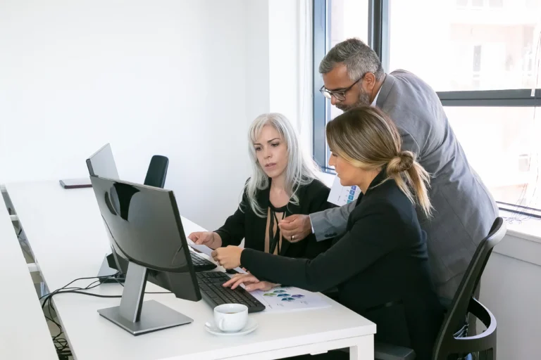 Business Team Three Analyzing Reports Sitting Workplace With Monitors Together Holding Reviewing Discussing Papers With Charts Copy Space Inclusive Workplace Concept